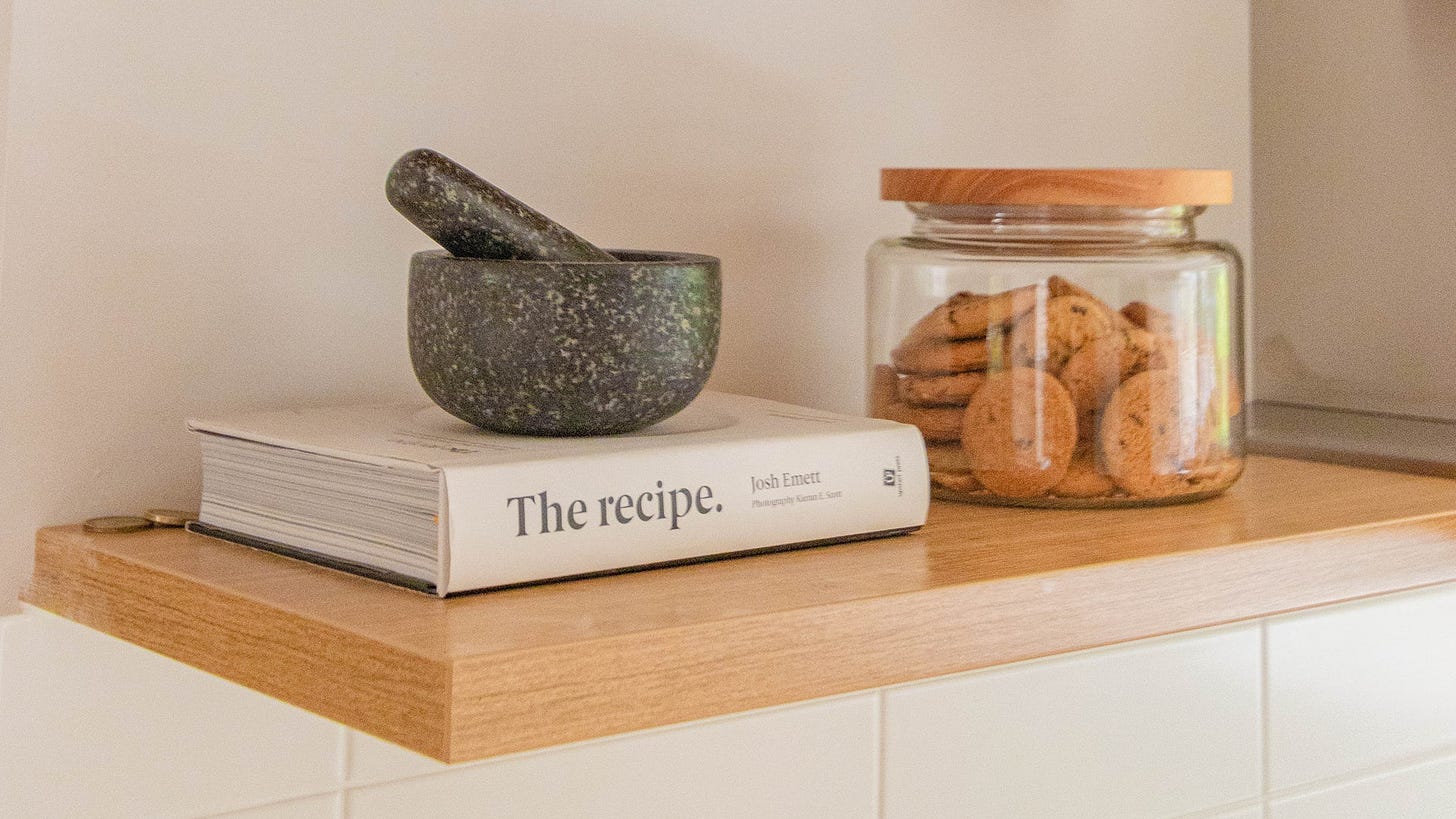 A photo of a kitchen shelf with a recipe book, mortar and pestle, and jar of cookies