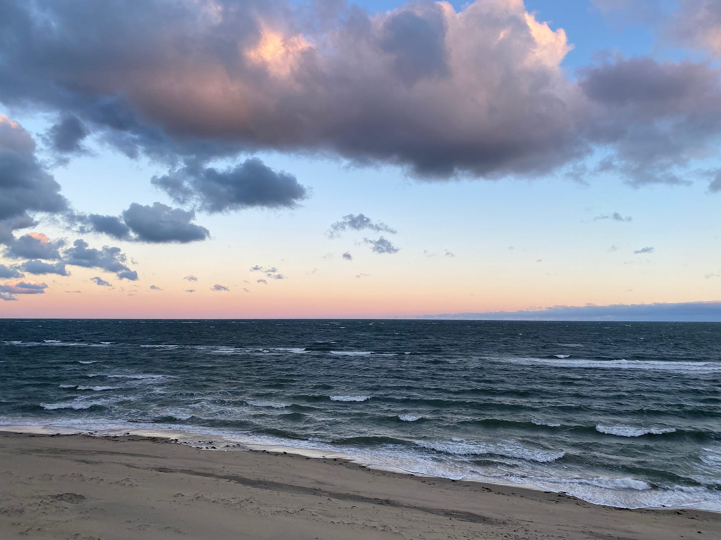 View of the beach at sunrise. The ocean is deep blue and ruffled with waves. The sky is pink on the horizon, then gold, then pale blue, then deep blue. There are big dark pink and grey clouds.