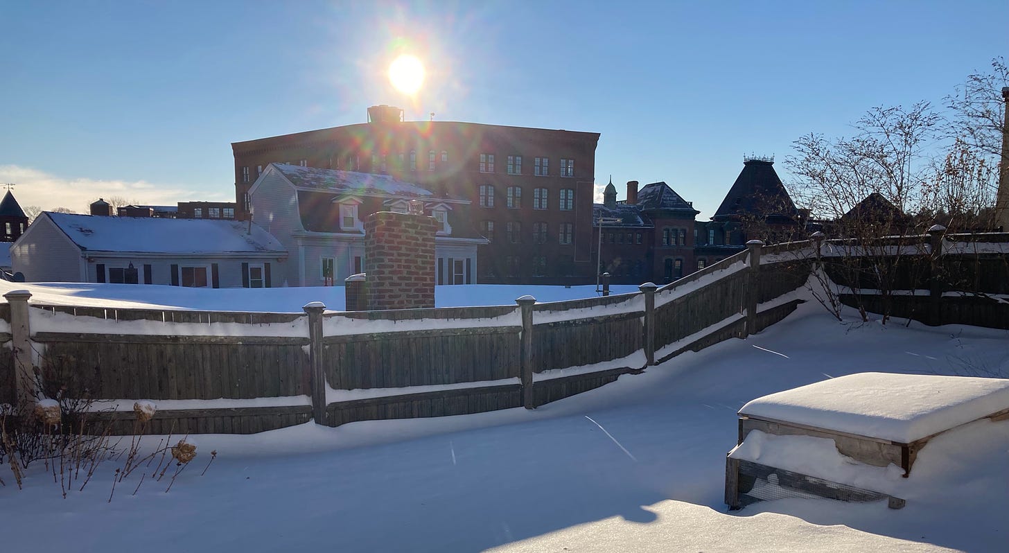 Deep, untouched snow in the foreground. A brick building in the background, with the sun rising just above it. The sky is a brilliant blue.