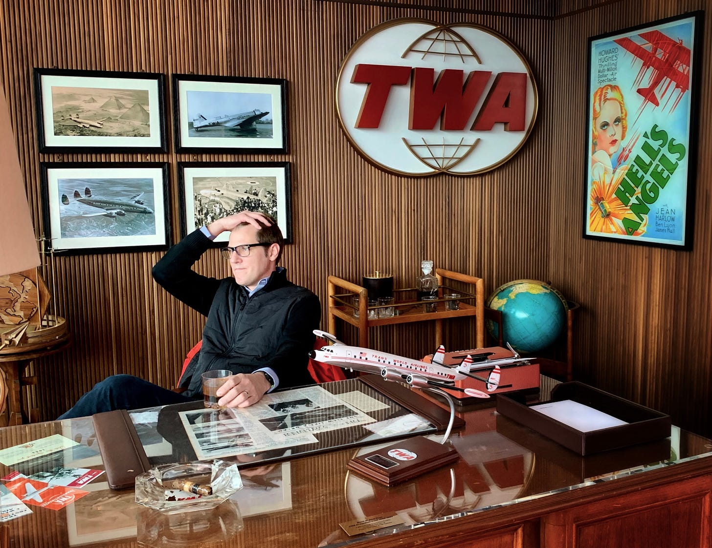 Tim sitting at a large oak desk on the set of a former TWA executive's office. He's holding one hand on his head, wondering something.