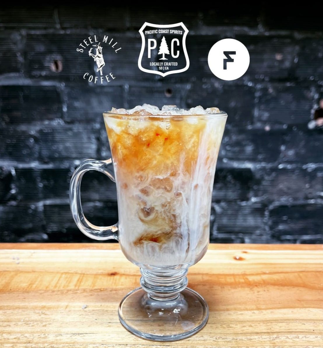 A tall, clear glass coffee cocktail with blended coffee and whiskey and cream on ice. The glass is on a wooden bar top in front of a black painted brick wall.