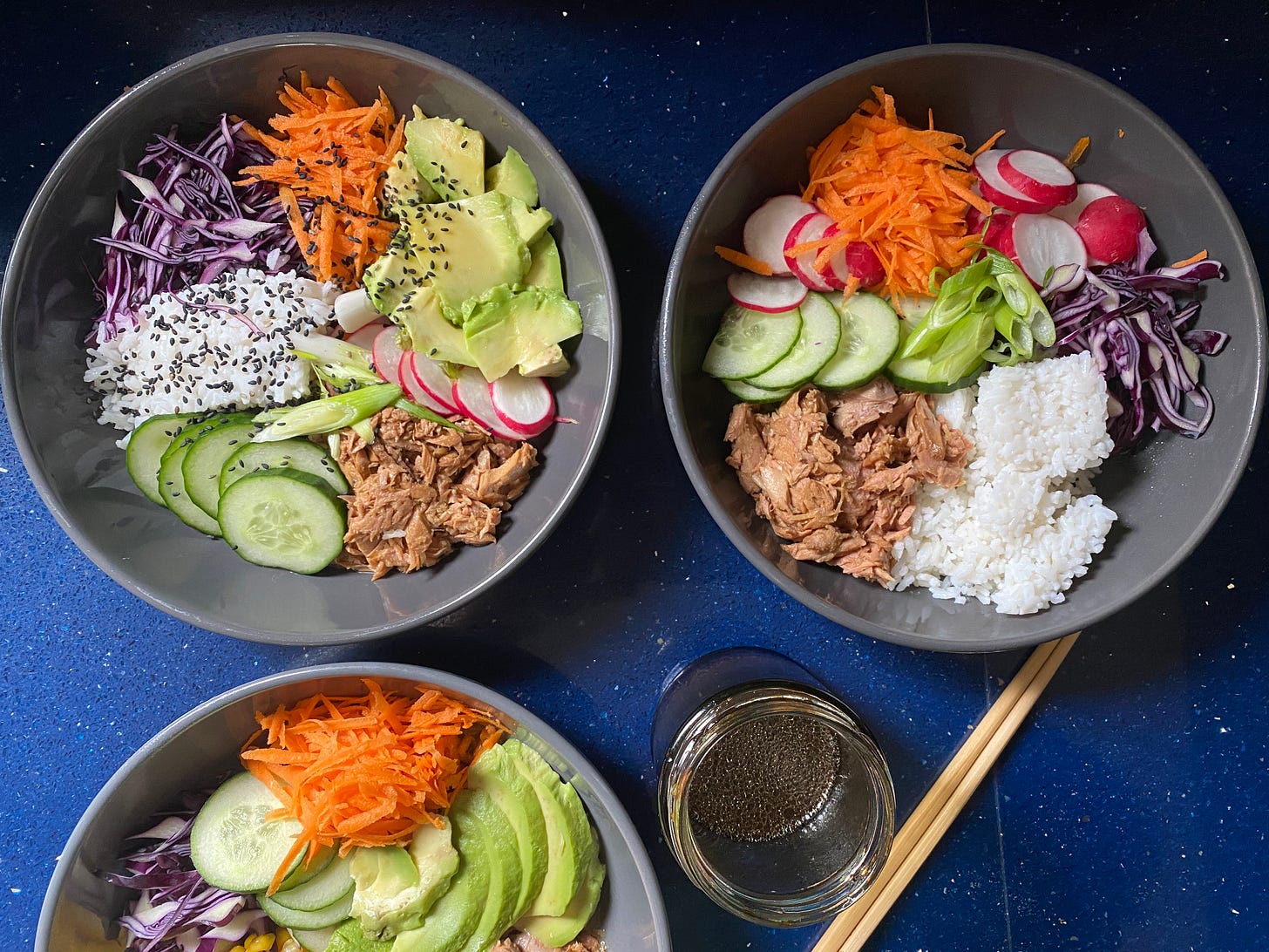 Three grey bowls on a blue quartz worktop. Each bowl is filled with chopped and shredded vegetables, some rice and tuna. A jar of sauce is next to them, and a set of chopsticks.