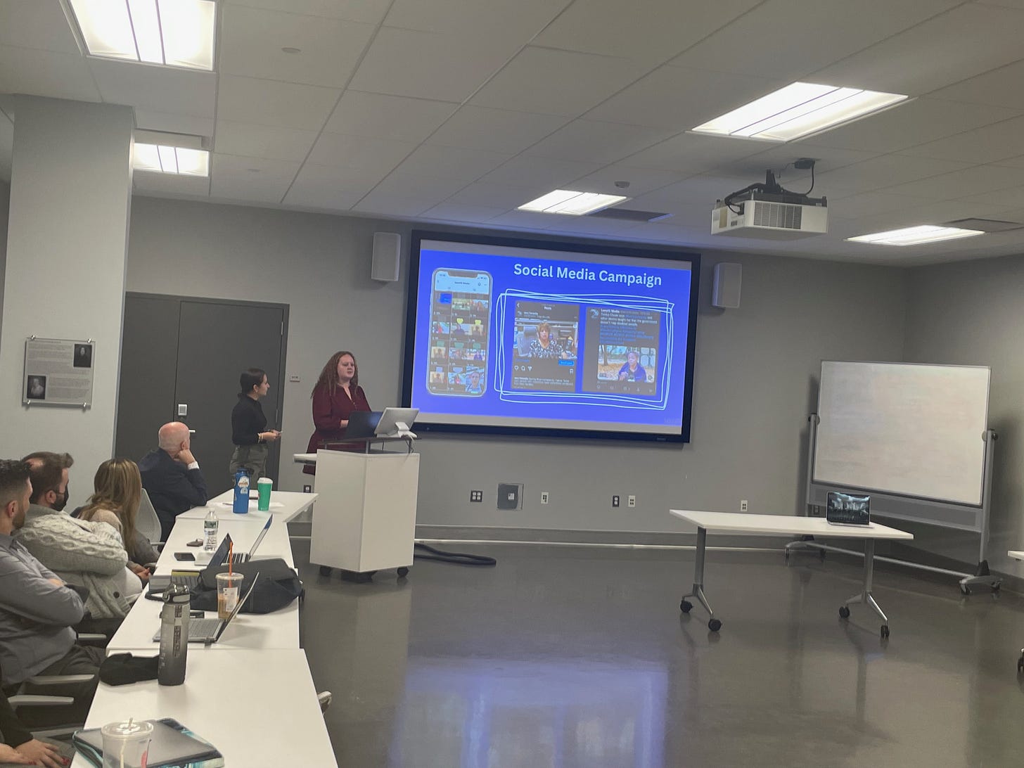 In a classroom, two women stand near a lectern adjacent to a projection screen. On the screen are the words social media campaign