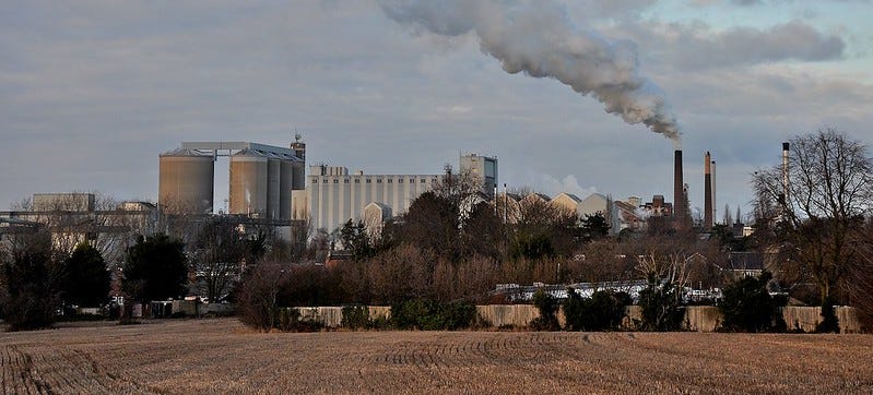 A panoramic photo of Bury St Eds sugar beet factory. Clouds of smoke gush from its chimneys. In the foreground are wintry fields used for wheat in the summer. 