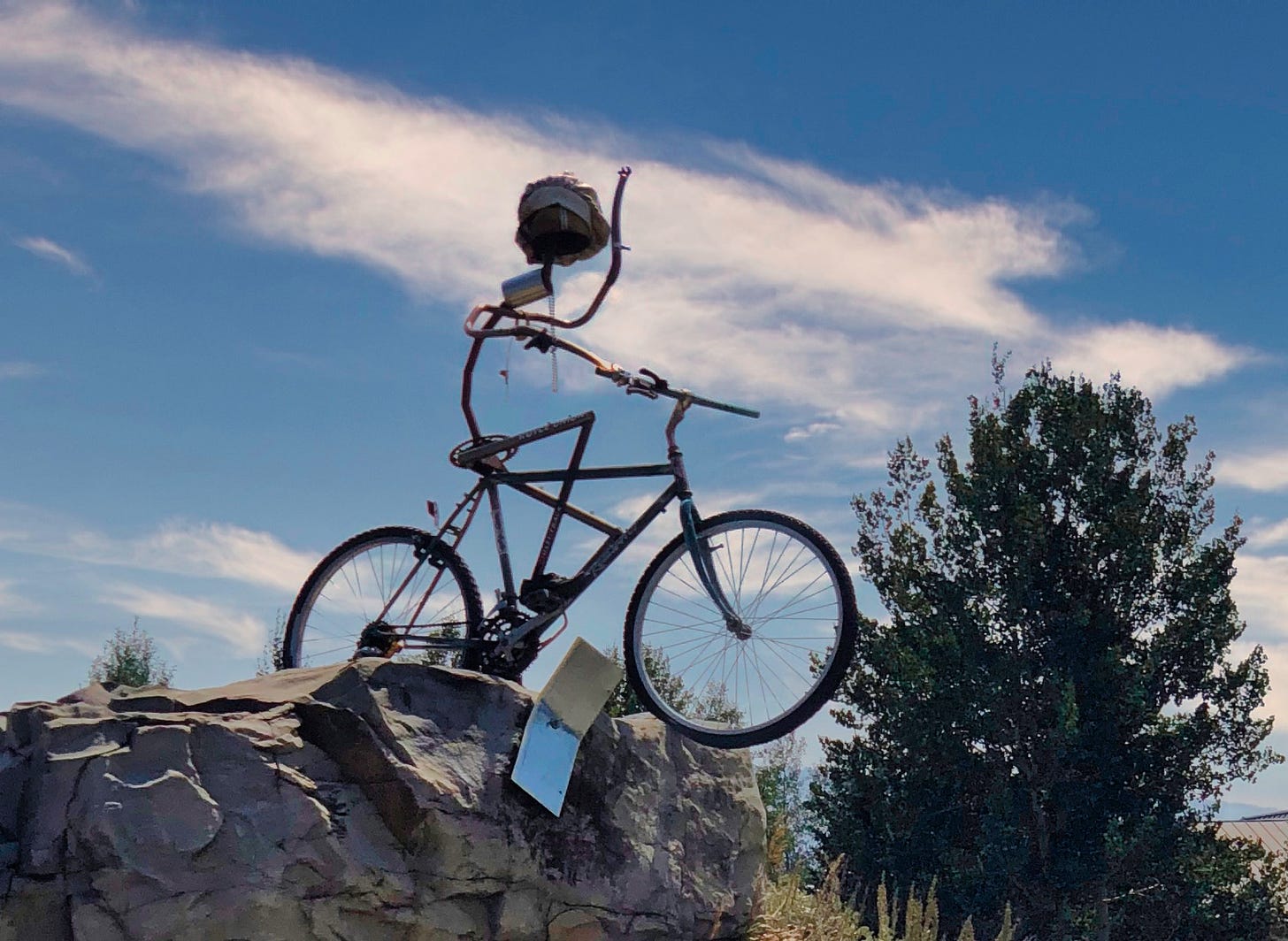 A sculpture of an old bicycle with a metal figure waving, wearing a white cap and about to ride off the side of a huge rock with their notebook falling near the rock and tire 