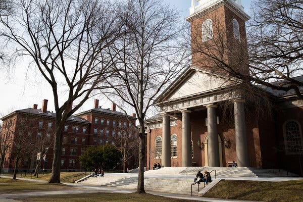 The Harvard Yard, with red brick buildings and trees bare. and students sitting on the steps. 