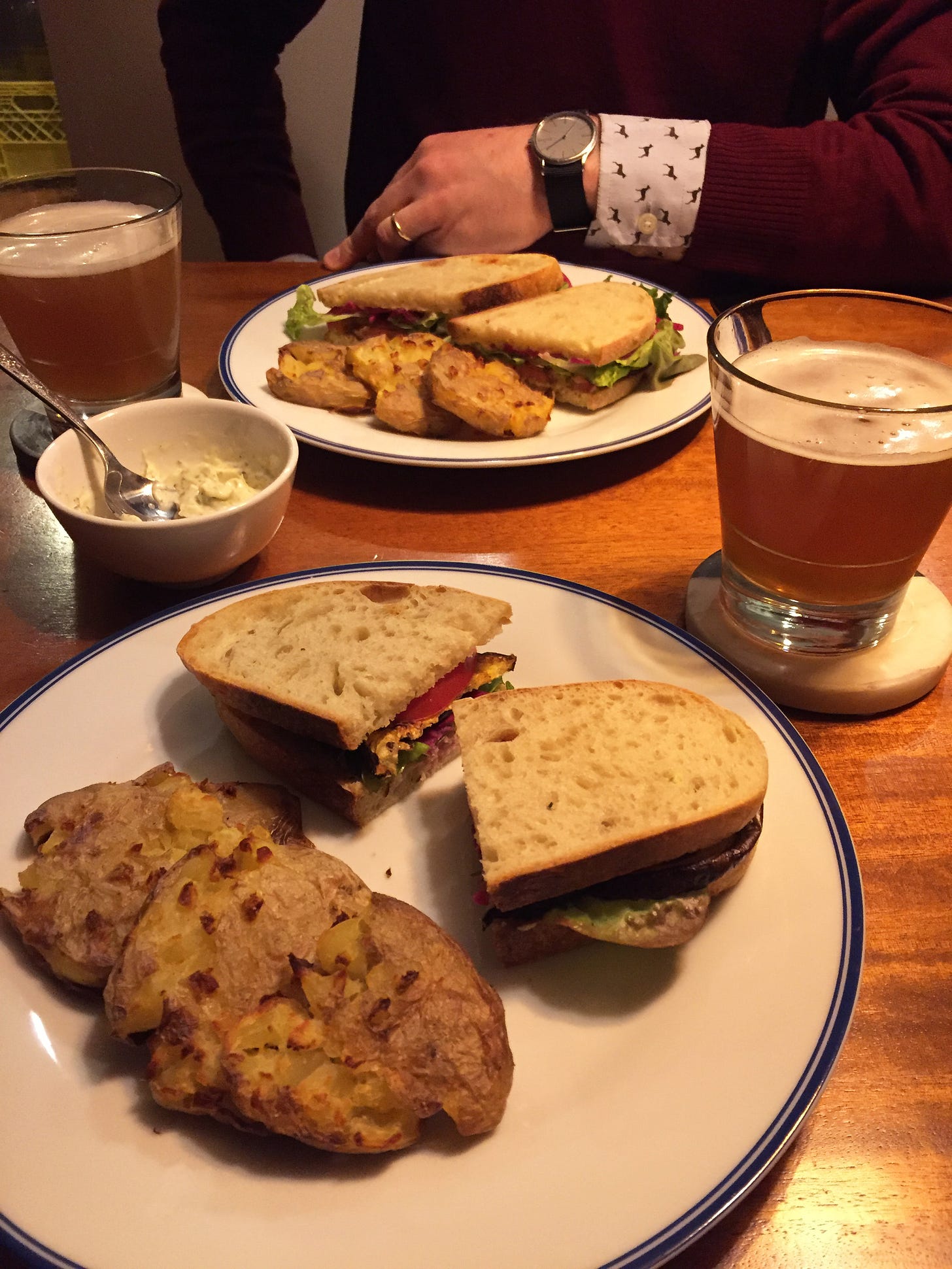 Two large plates with sandwiches cut in half and three smashed potatoes arranged next to them. Between the plates is a small dish of basil mayo, and two glasses of beer on coasters sit to the sides of the plates.
