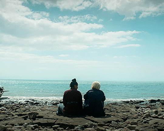 This GIF (repeated moving image) shows Yaz Khan (played by Mandip Gill) and the Thirteenth Doctor (played by Jodie Whittaker) sitting on a Chinese beach in the year 1807 while talking to one another. Yaz sits on the left, close to the Doctor, who sits on the right. Both have their backs to the camera, while the beautiful blue ocean and sky are visible beyond them.