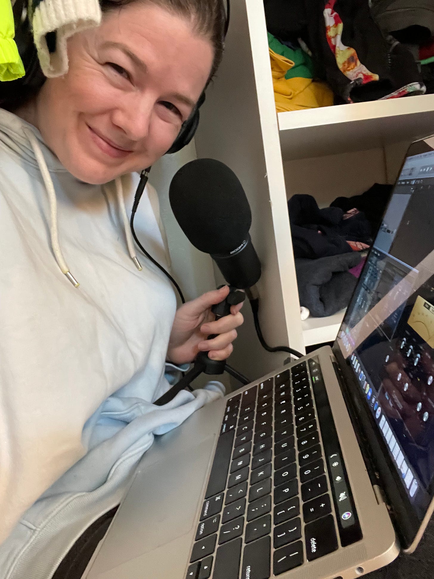 A white woman in a light blue sweatshirt peaks out from a closet with headphones, a microphone and a laptop. 