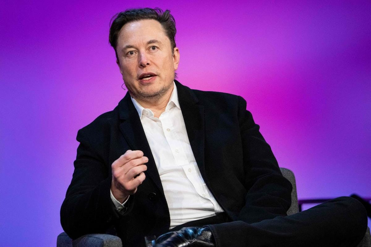 Elon Musk sits in a chair with one ankle crossed over the opposite knee. His mouth is open and one of his hands is slightly raised.