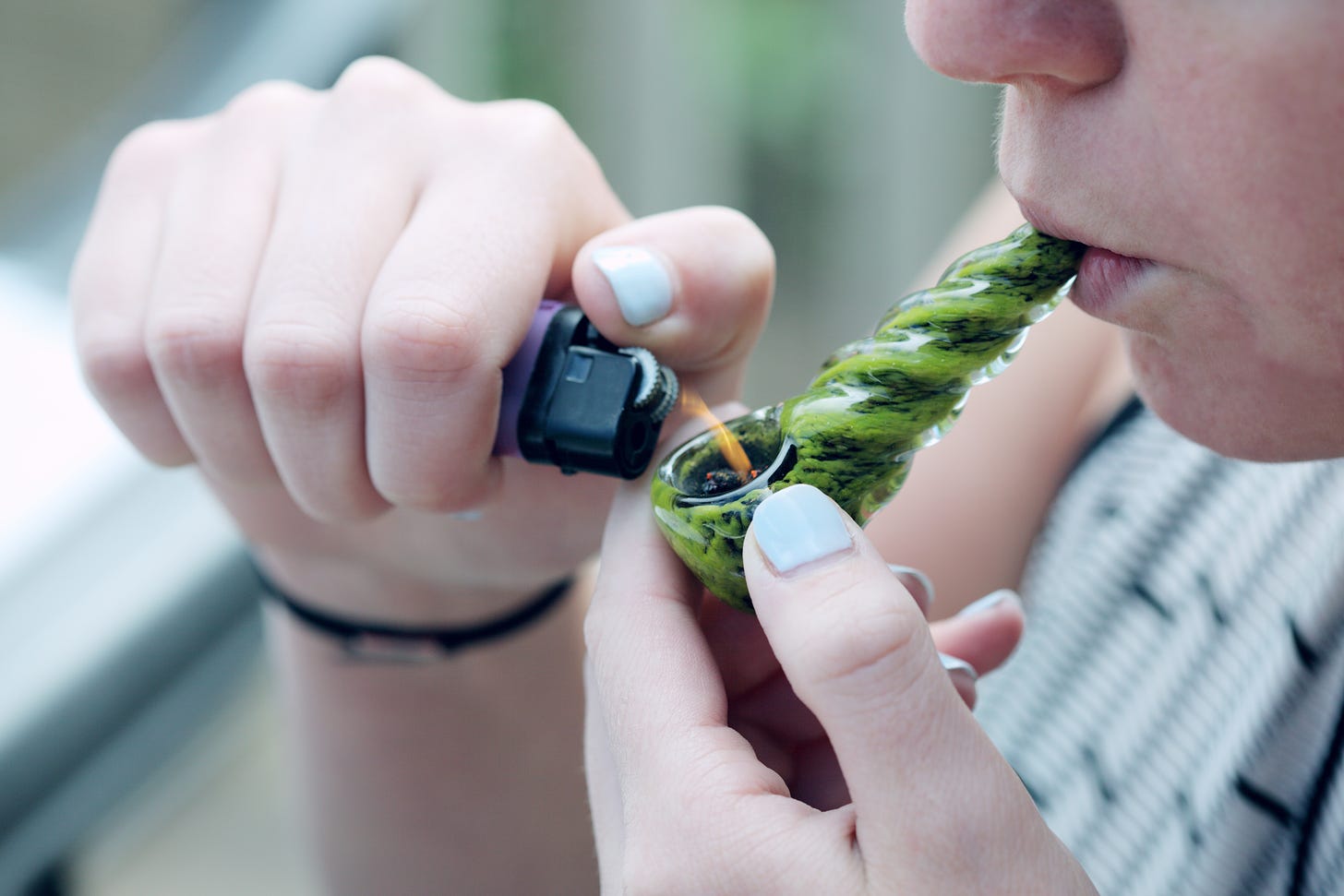 person with light blue nail polish lighting the bowl of a green glass pipe with marijuana in it