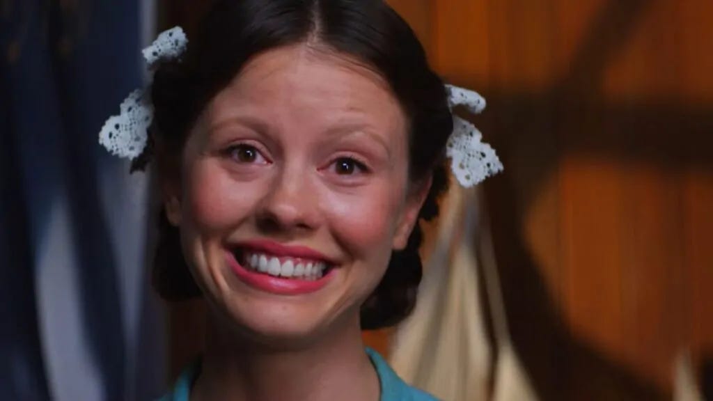 Print de tela do filme Pearl, em que a atriz Mia Goth, caracterizada como a protagonista do filme, está centralizada na imagem com o rosto congelado em um sorriso extremamente exagerado, com lágrimas nos olhos e bochechas avermelhadas. Ao invés de feliz, ela parece desesperada.