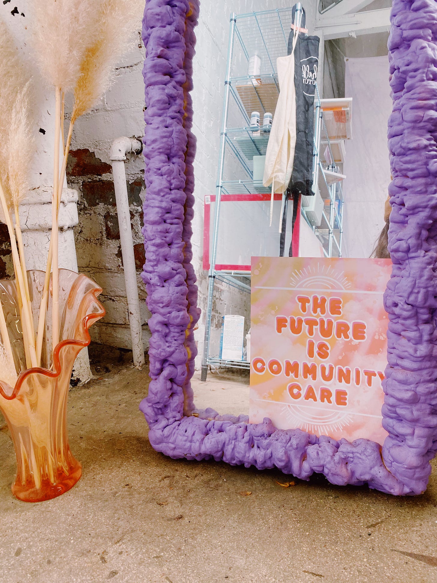 Photo of The Future is Community Care art print in a mirror with lilac foam frame on a concrete floor next to a pink glass vase with pampas grass. 