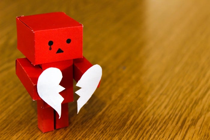 A paper mache robot with a tear under one eye holding a broken heart.