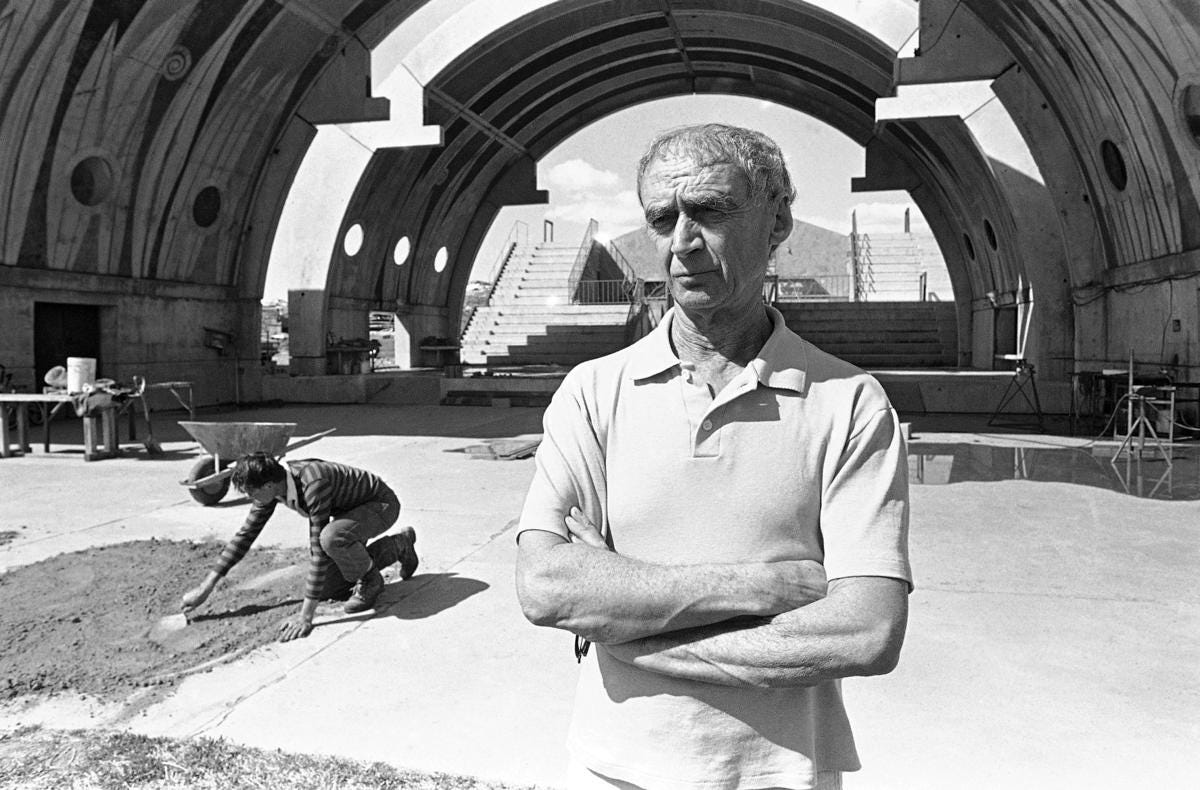 Paolo Soleri stand Italianly, arms folded, as behind him a student on hands and knees lays concrete at Arcosanti