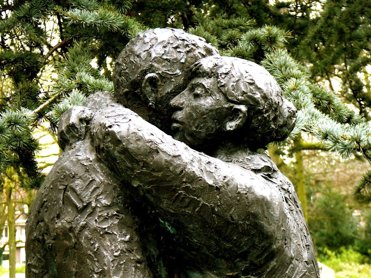two mottled gray statues are hugging each other in a deep and meaningful way while there are pine trees in the background