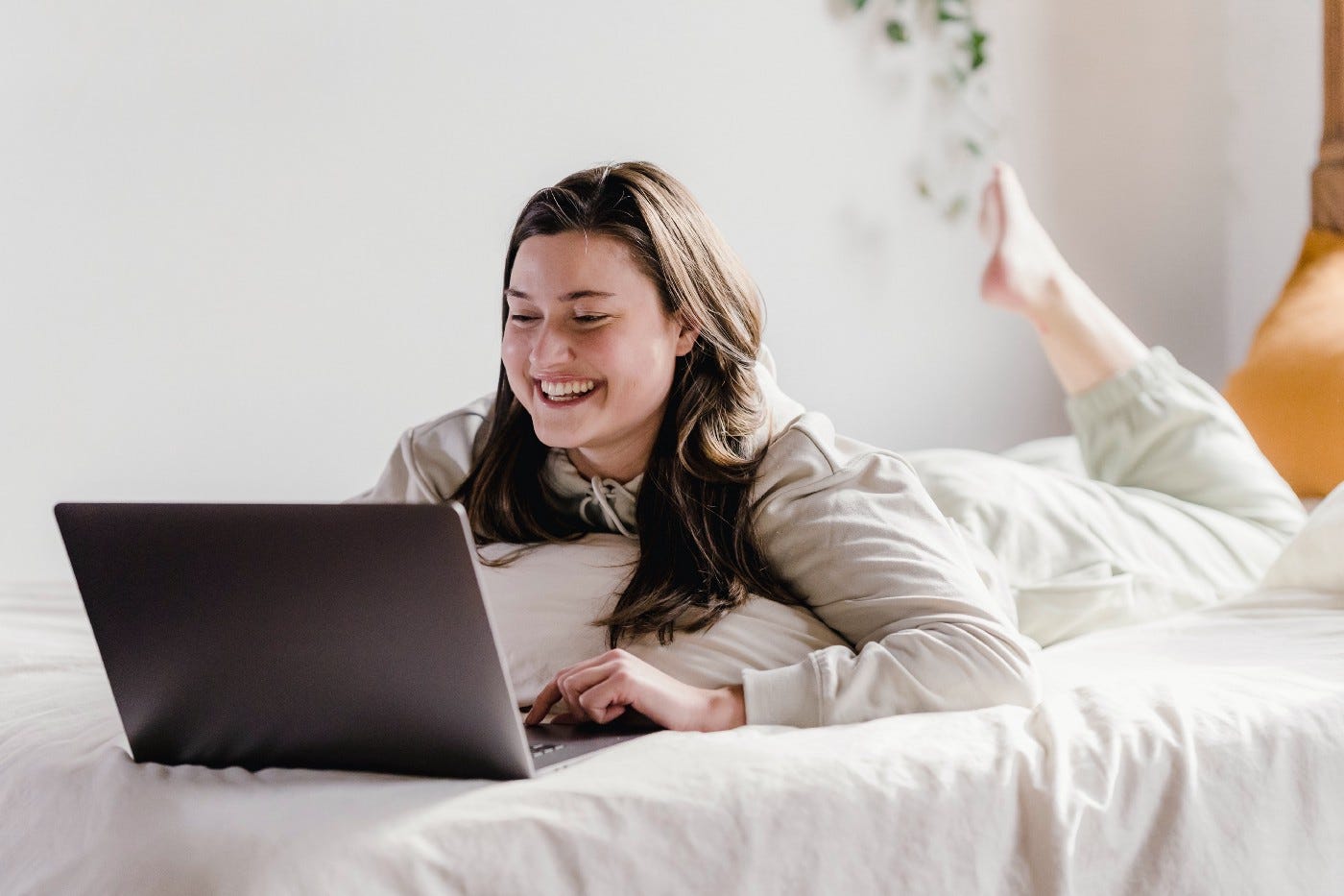 Woman laying on a bed, working on her laptop. She has one foot in the air. She is smiling. Bed and background are white and she is wearing white. Very minimalistic photograph.