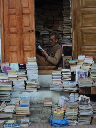 Loyal Reader #4 Erin taught at an elementary school in Morocco for two years. Here’s her favorite bookseller inside his used bookstore stall in the Medina of Rabat. (Erin reports that selling books wasn’t his top priority.)