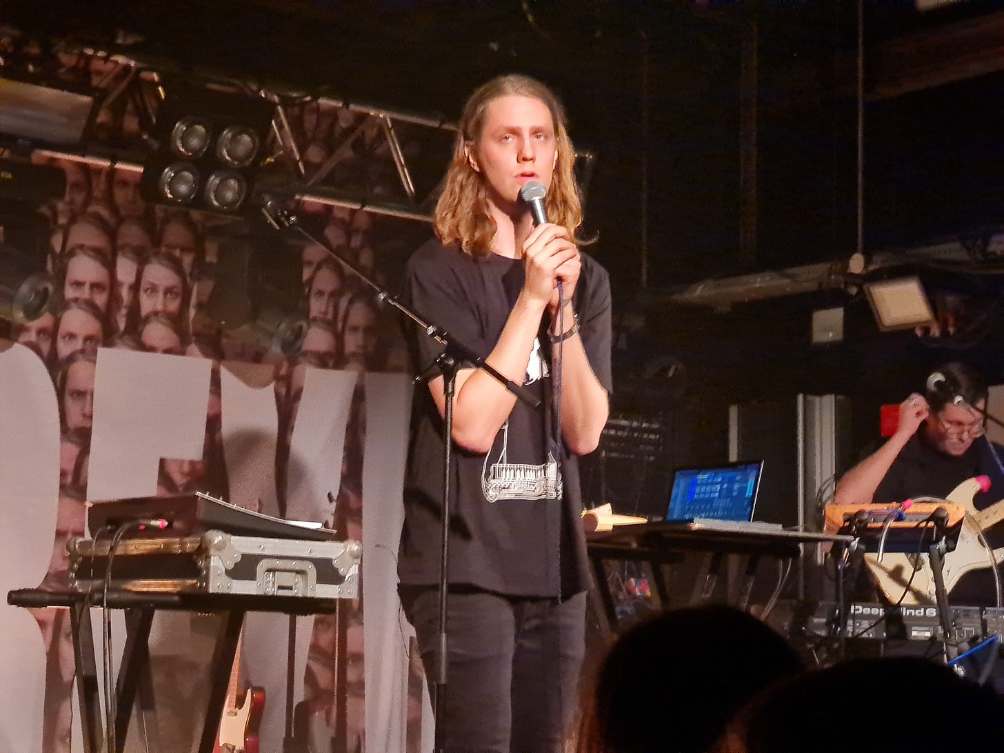 Daði Freyr, a tall white man with long blonde hair, is holding a microphone and looking at the camera. He is surrounded by electronic instruments and a laptop, and there is a dark-haired man playing guitar in the background.