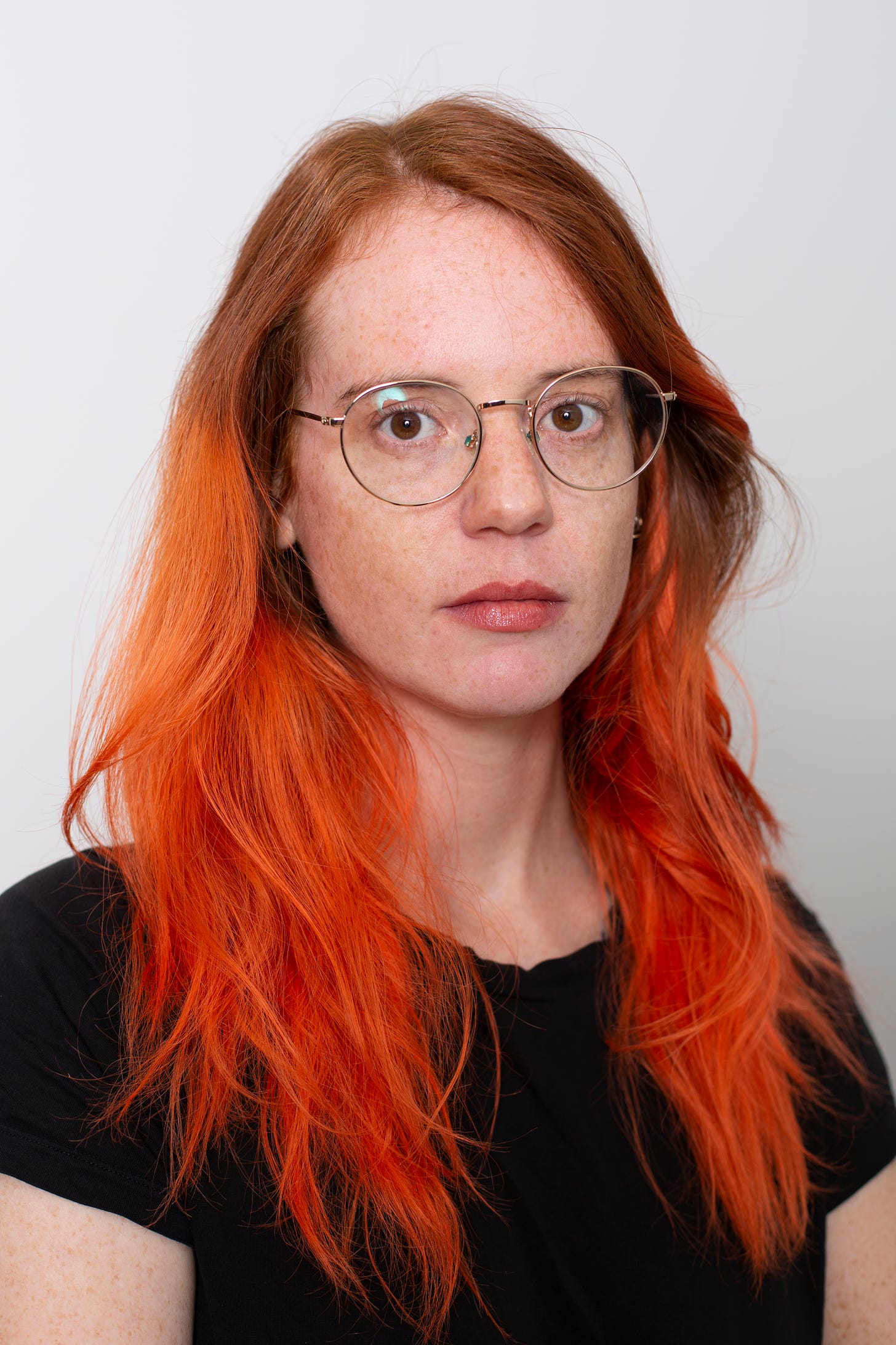Photo of Beatrice Adler-Bolton, portrait of a white woman with natural red to dyed orange long wavy hair and freckles, wearing gold round-rimmed glasses and a black t shirt in front of a white background.