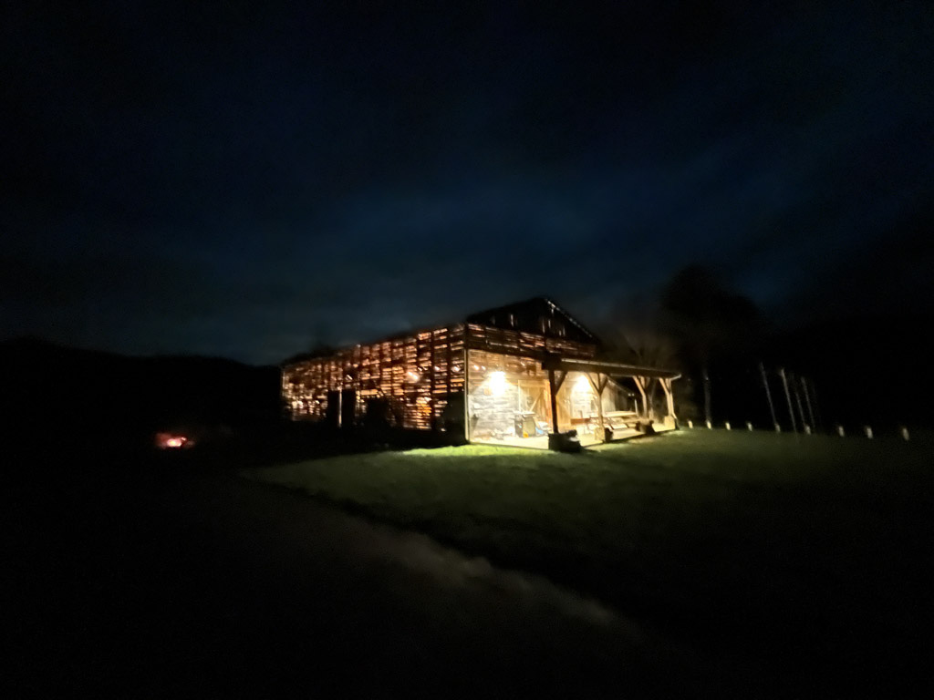 A soft, dark photograph of a tobacco barn in a field at night. Bright light escapes through the siding.