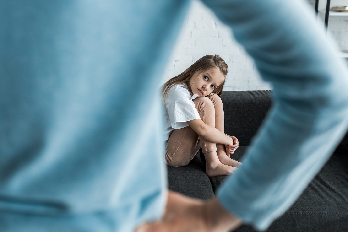 A child cowers on the coach and looks anxiously up at their mother who has her hands on her hips.