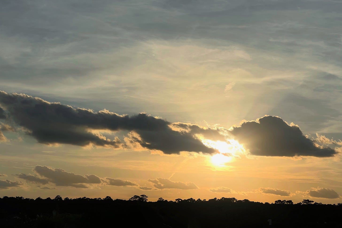 Sunset with the bright sun peeking behind grey clouds shooting rays into the buish clouds above and toward the trees in the horizon below