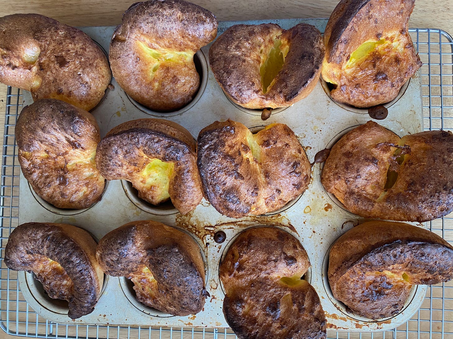 A muffin tin holding 12 tall golden brown popovers, all puffed up in various irregular shapes, most with a small crack in the middle showing the lighter golden inside.