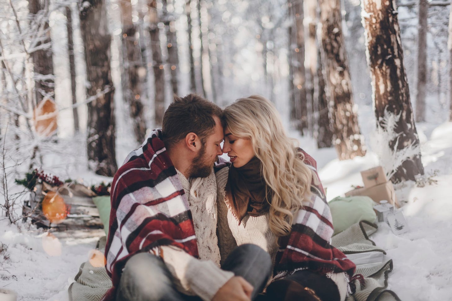 A couple sits in a wintery wood with with their heads pressed lovingly together.
