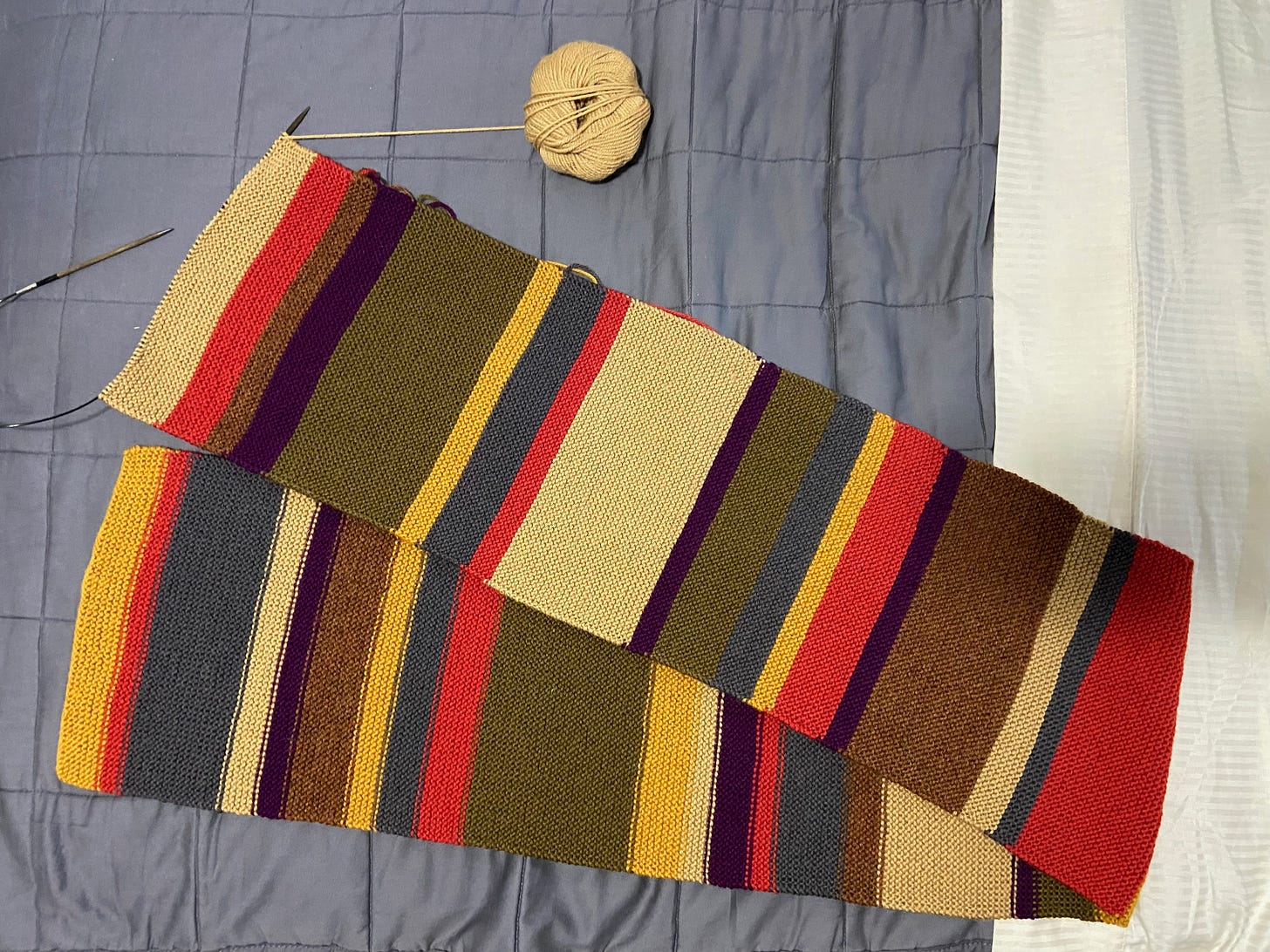 An in-progress DOCTOR WHO scarf displayed on a bed. The most recent color attached to the needles (and with a ball of yarn off to the side) is tan.