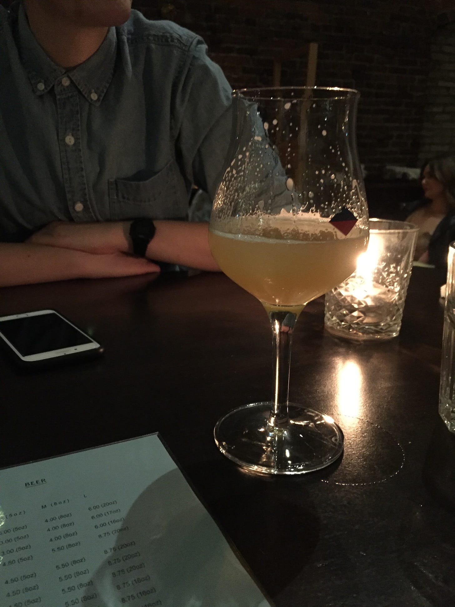 At a dark pub table lit by a candle, a bell-shaped glass of pale beer sits next to a beer list. In the background, there is an iphone on the table and a person in a button-up shirt whose face is not visible.