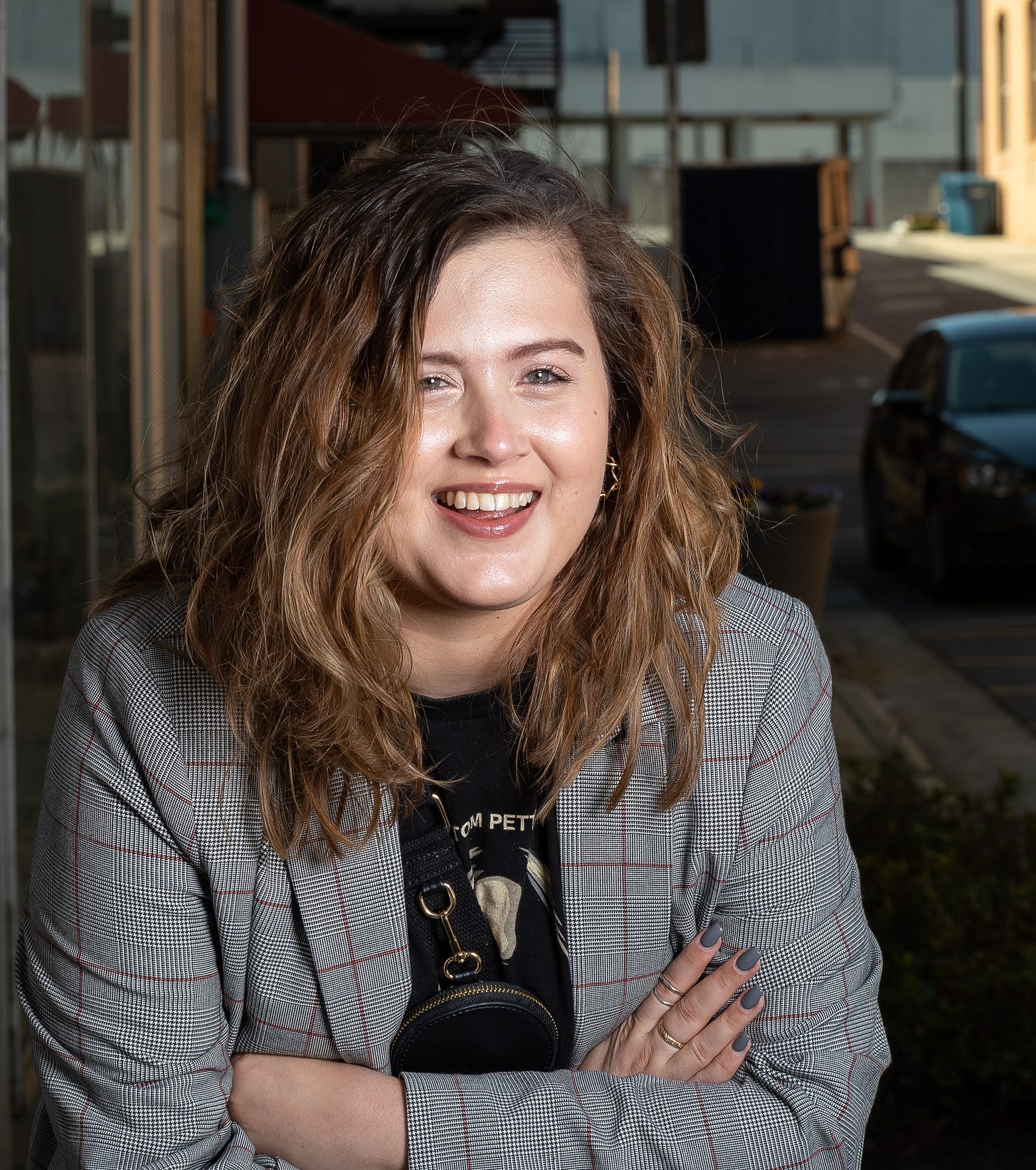 Rikki, a smiling woman with shoulder-length brown hair smiles for the camera while crossing her arms. She wears a blazer with a Tom Petty shirt.