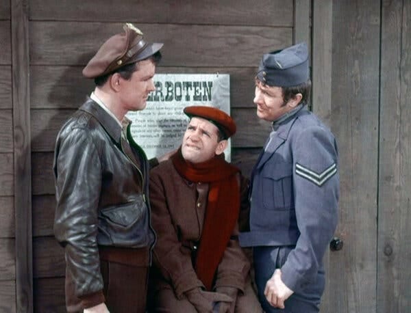 Robert Clary, wearing a red beret and a matching scarf, standing in front of a wall with a sign that says “Verboten” between two men, one in a leather jacket and cap and the other in a military uniform.