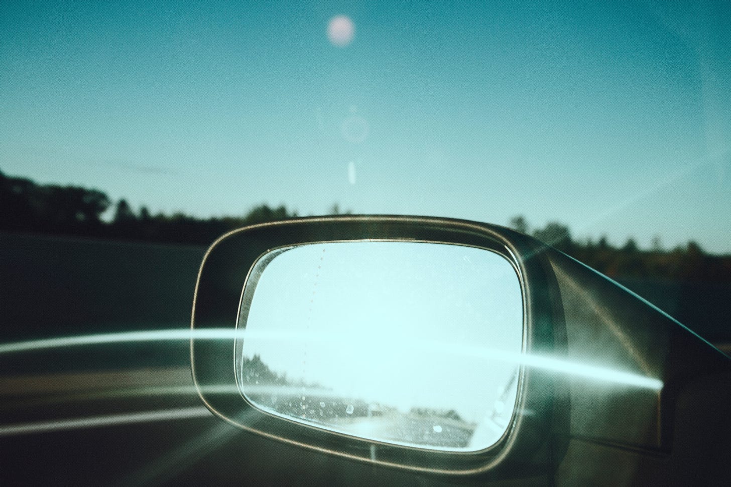 A car's side view mirror with the sun glinting off of it, against a blue sky and an obscured view of a highway.