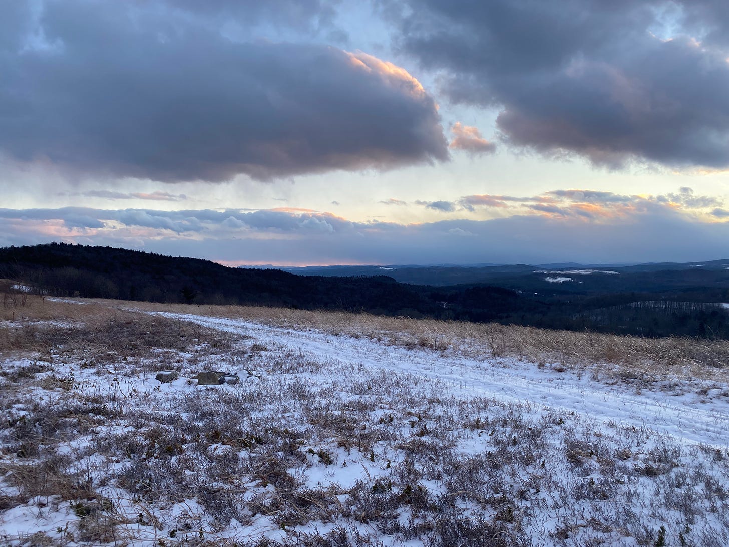 View of a snowy hill at sunset, looking out over an expanse of hills and fields. The sky is full of dark clouds edged in gold.