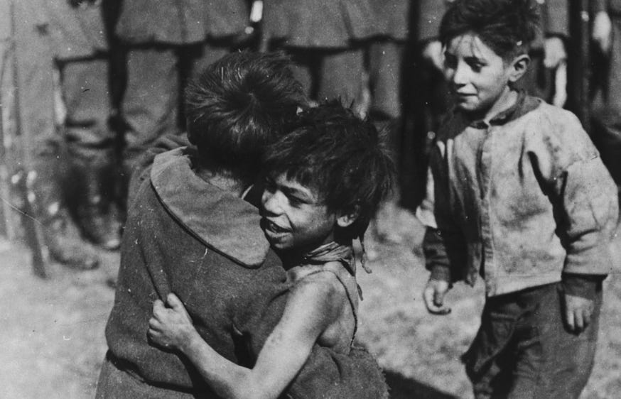 Image description: A black and white original photograph of Romani children in a concentration camp during the Holocaust. Two of the children look like they have tan skin tones and are hugging each other with one of them facing the camera and smiling, while another lighter-skinned child looks on, smiling. They are dressed in poor and dirty seeming clothes. Behind them stand others, possibly adults, but only their legs are visible so it’s not clear. The people in the background are wearing boots