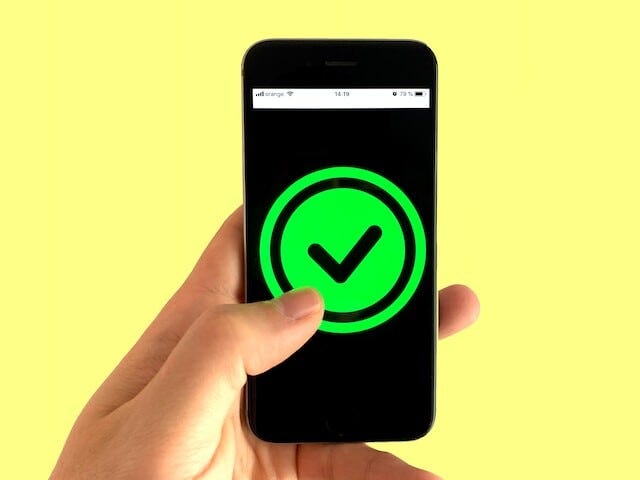 hand holding a phone against a yellow background. On the phone screen there is a green circle with a black check mark inside it