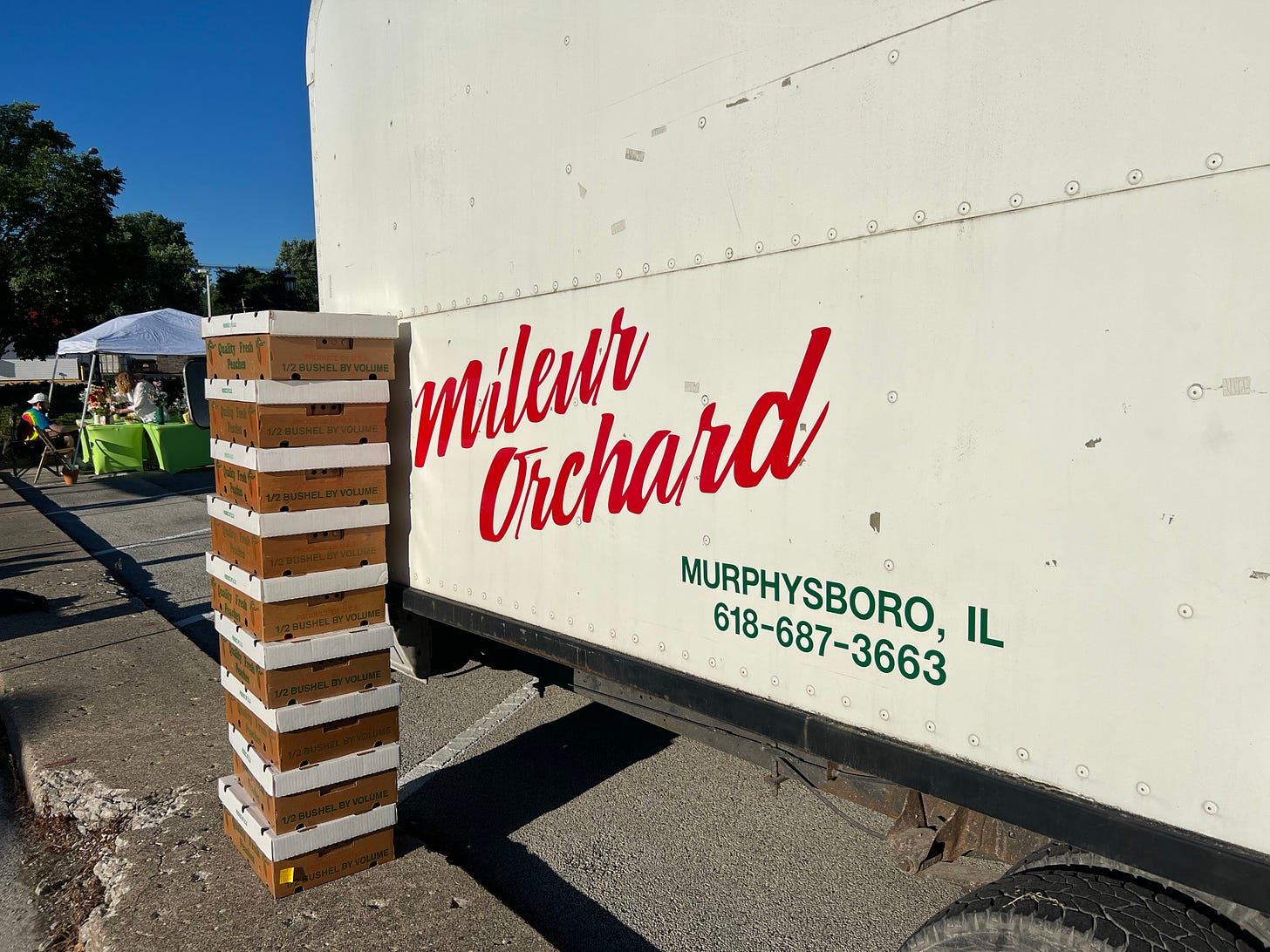 A stack of peach boxes against a truck that says Mileur Orchard Murphysboro, IL 618-687-3663