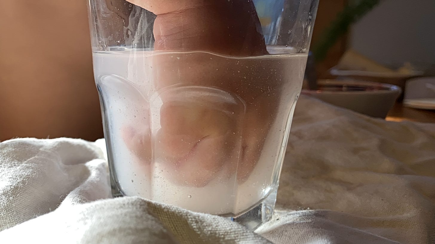 A toddler's chubby hand submerged in a glass filled with cloudy liquid.