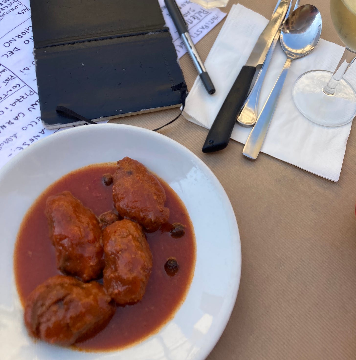 A notebook, a glass of wine, a plate of sardine balls in tomato sauce