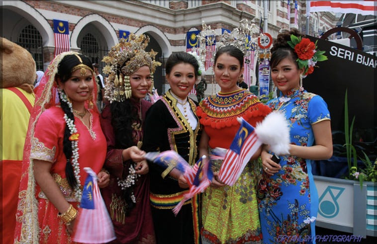 A picture of 5 women each wearing the traditional dress of their ethnic background. They are all holding the Malaysian flag. Woman on far left is wearing a type of Indian dress that is a pink red color. Woman on far right is wearing a type of Chinese dress that is a light blue. The three women in the middle are wearing different types of Malay dress that are red and yellow with silver headdress, black and white with red/yellow/gold stripes, and dark burgundy red with gold headdress respectively.