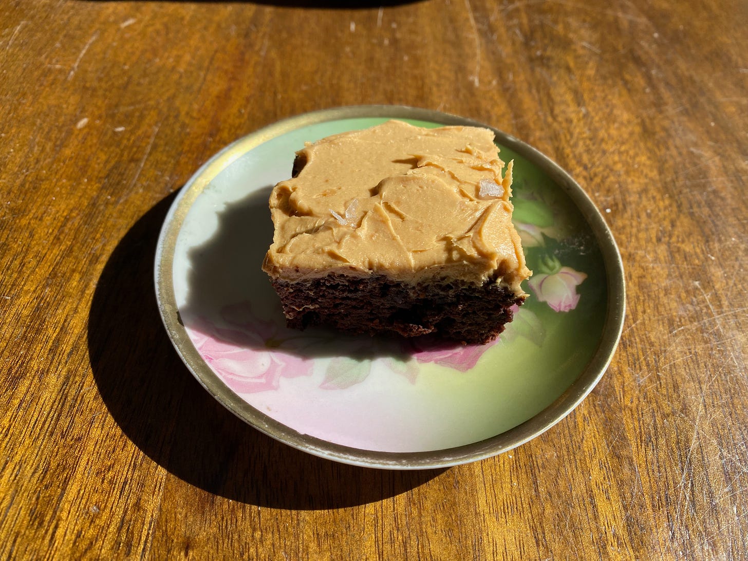 A brownie topped with peanut butter frosting and flecked with flaky salt sits on a small plate painted with roses on a wooden table.