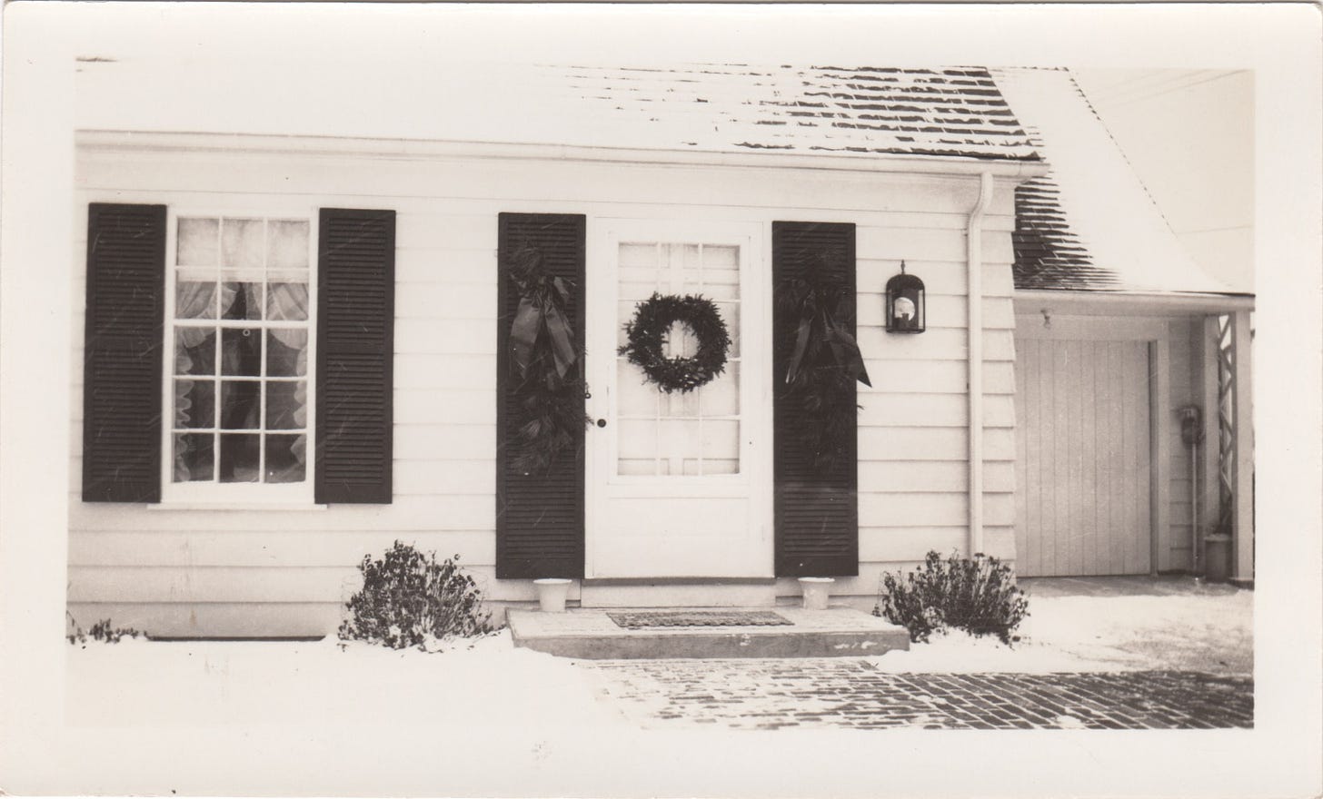 Photo of a White House with dark shutters on either side of a front door and adjacent window. On the door is a wreath, and on each shutter, a large evergreen spray.