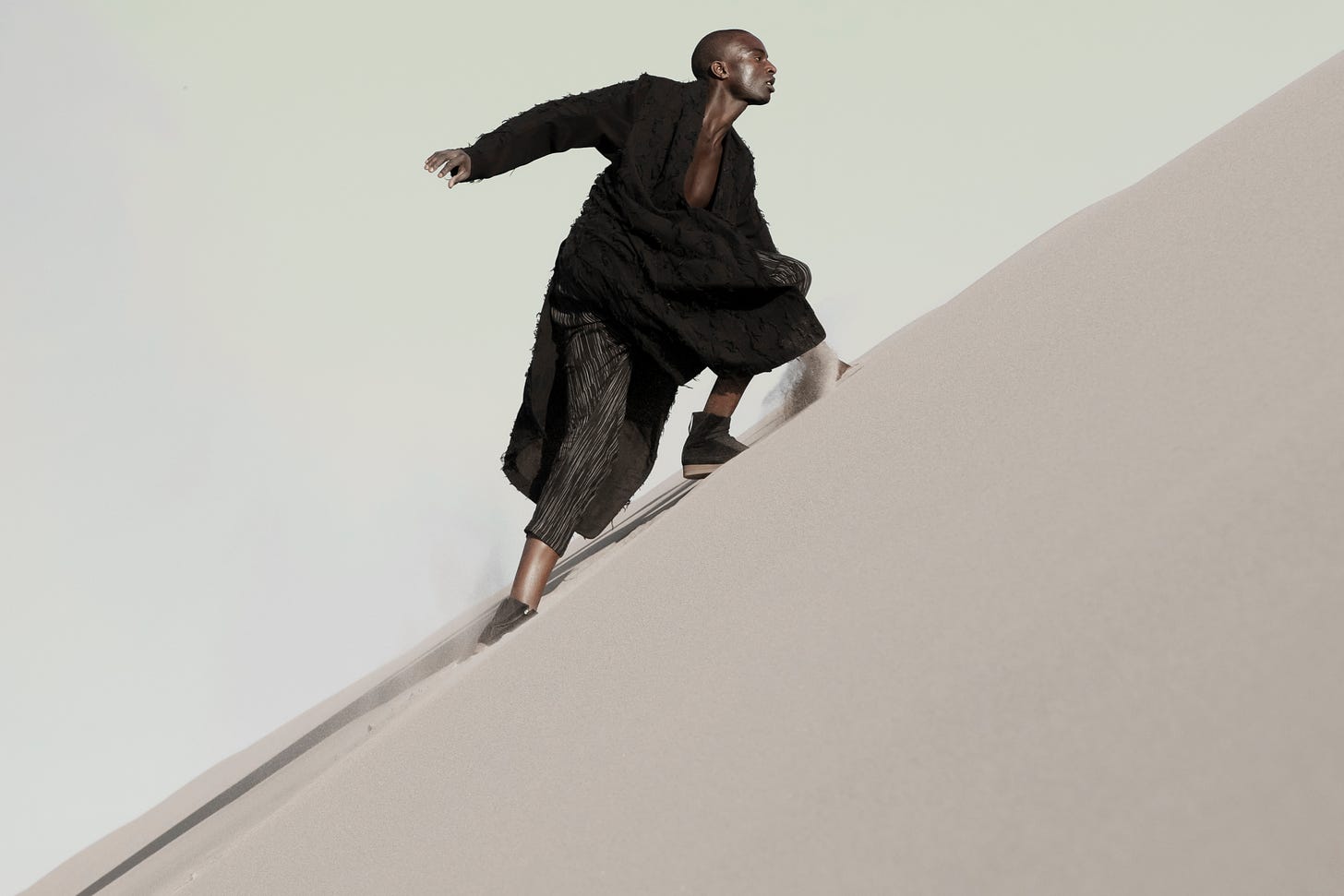 A model wearing a loose black jacket and trousers, dashing up a slope of desert sand. Photograph by Rob Daly for Abasi Rosborough