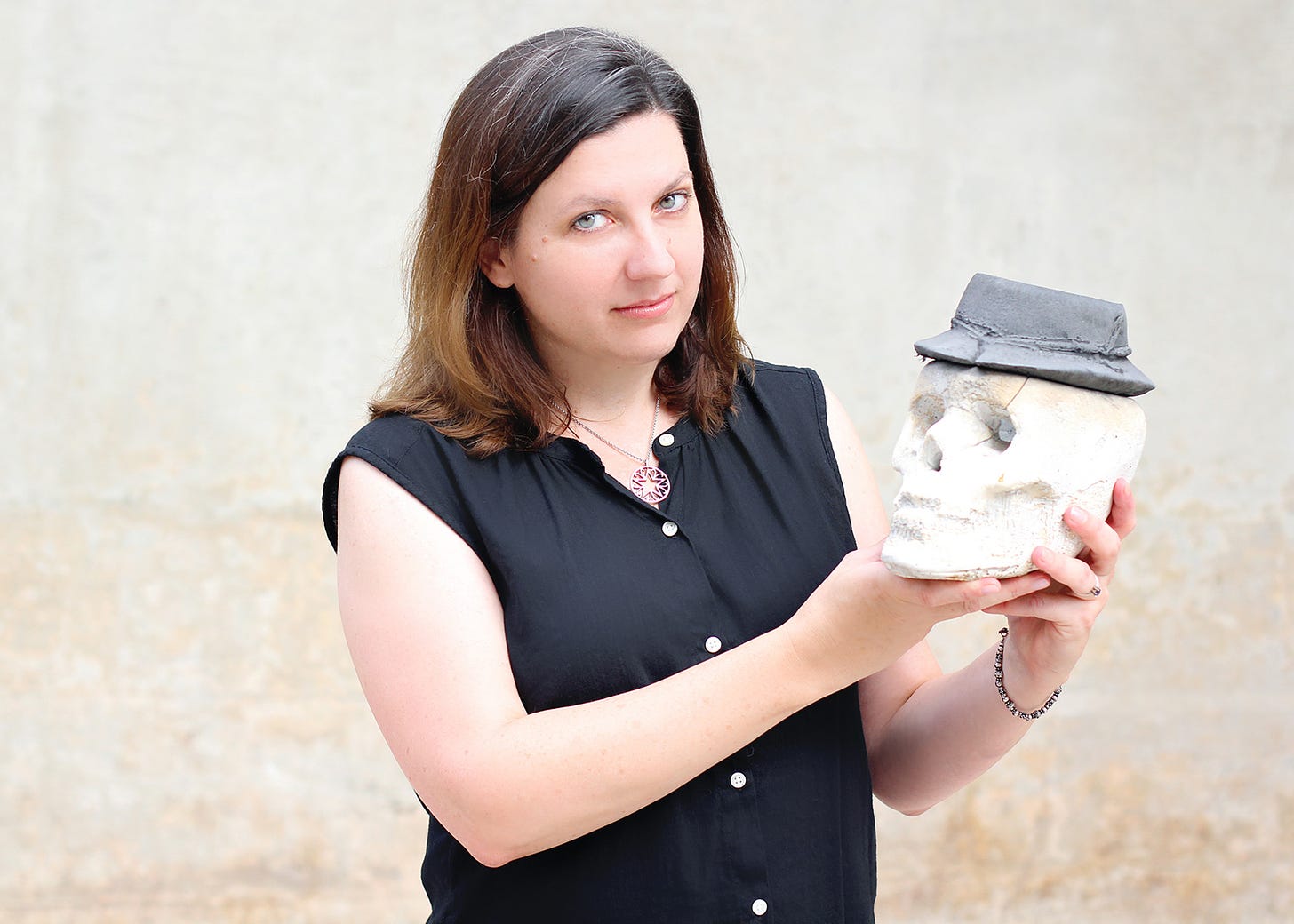 A woman in a black shirt looks at the camera. She holds a white ceramic skull with a black hat. 