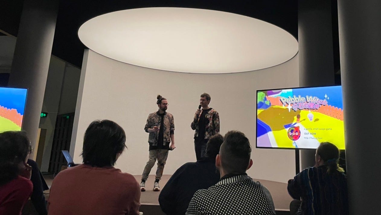 Pete and Scott standing on stage in front of an audience at Powerhouse Museum. One of their games "Pebble Witch" is displayed on a large screen beside them.