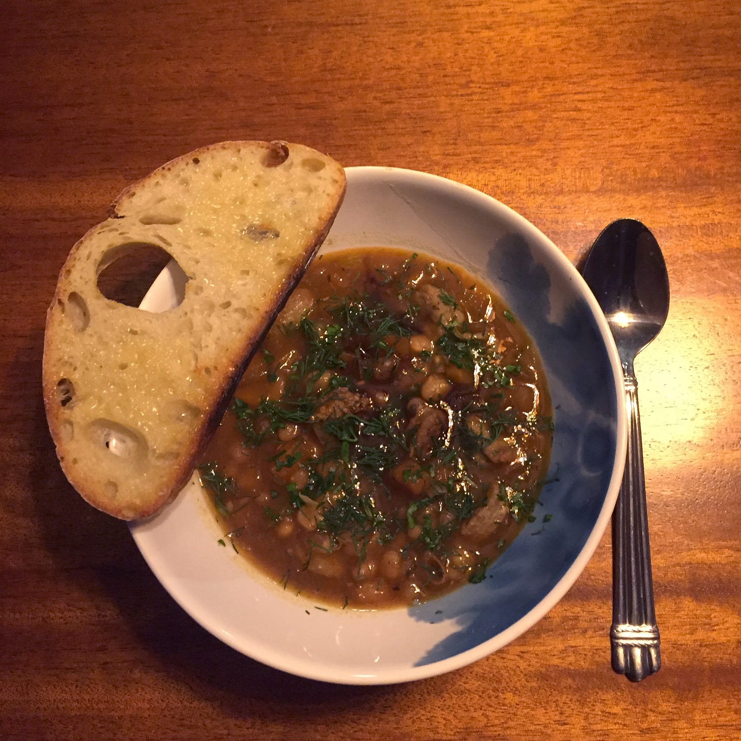 From above, a bowl of bean stew sprinkled with herbs, with a slice of sourdough resting on the edge of the bowl and a spoon on the table beside it.
