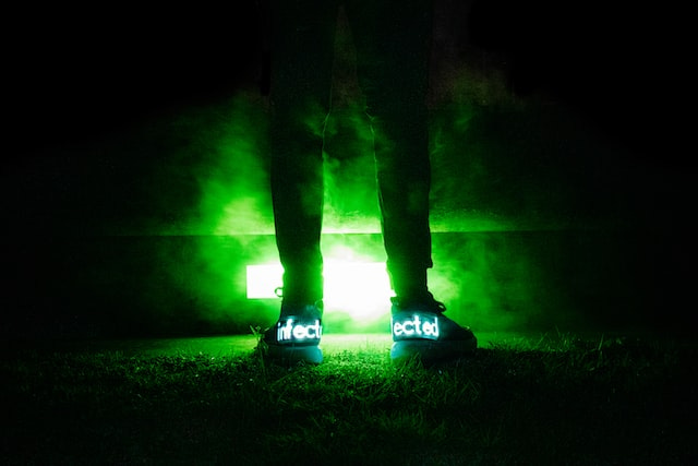 Black background. Two sneaker clad legs are in silhouette by the light of an acid green light. The word infected is illuminated on the shoe.