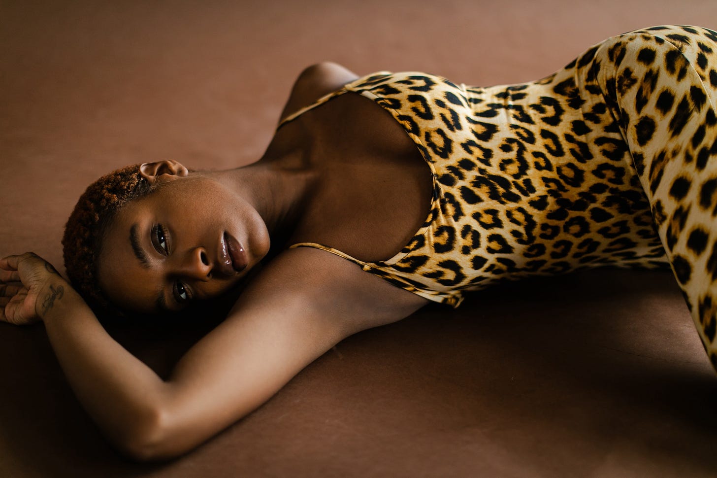 Attractive black woman with short-cropped hair reclining senusuously against a brown background in sensuous leopard tights