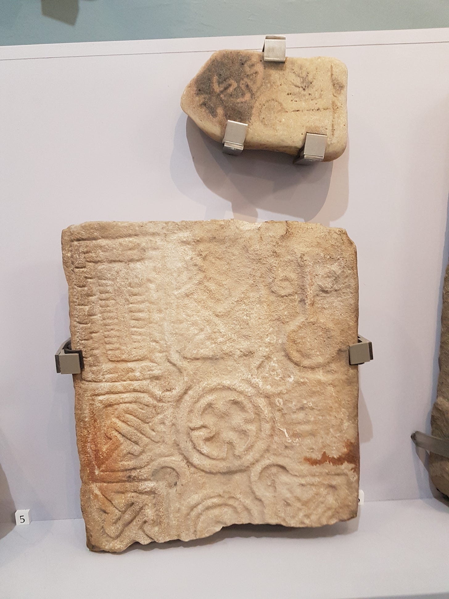 The Kinloss Stone and Drainie 32 displayed together in Elgin Museum. Drainie 32 is larger and has a cross head with interlace-filled arms and a Maltese cross-style roundel in the centre. Pictish mirror and comb symbols flank the top arm of the cross. There is key pattern decoration flanking the shaft of the cross. The Kinloss Stone is a smaller fragment,, with a similar Maltese cross-style roundel and more decoration that is quite hard to make out. (Accession Number ELGNM 2019.10)